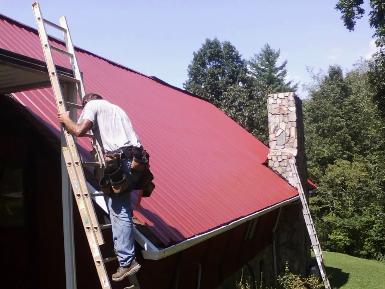 Roofer on a ladder beside a red metal roof and stone chimney — Soffit & Fascia Repair by Roofing Brothers in Palos Hills